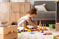 Toddlers playing with Montessori wooden toys such as stacking rings, puzzles, and pull toys in a bright, sunlit playroom with wooden shelves.